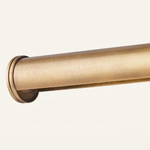 May include: A cylindrical curtain rod with a brushed brass finish and a smooth, rounded end cap. The metal surface features a subtle, muted gold tone with a soft, matte texture that extends horizontally across the frame of the image.