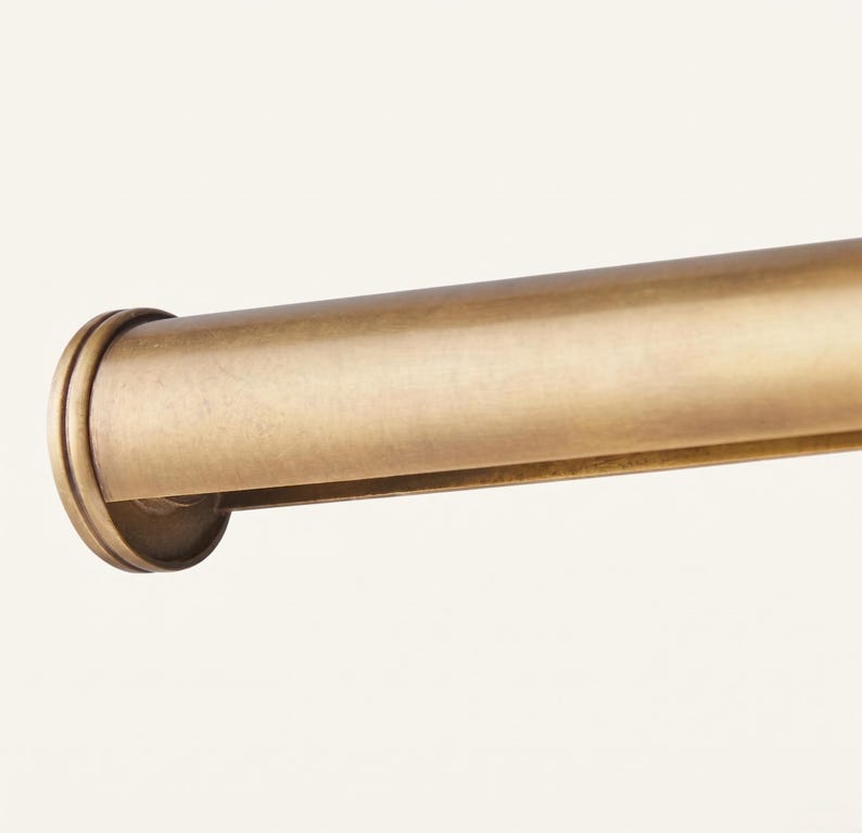 May include: A cylindrical curtain rod with a brushed brass finish and a smooth, rounded end cap. The metal surface features a subtle, muted gold tone with a soft, matte texture that extends horizontally across the frame of the image.