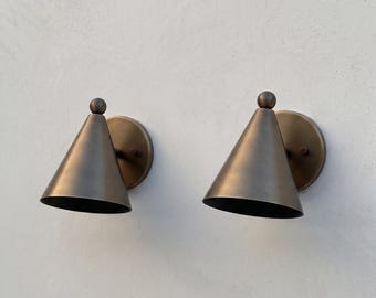 May include: A pair of wall-mounted sconces featuring a conical shade design in a brushed brass finish. Each light fixture consists of a circular backplate, a downward-angled cone-shaped shade, and a small spherical finial at the top apex.