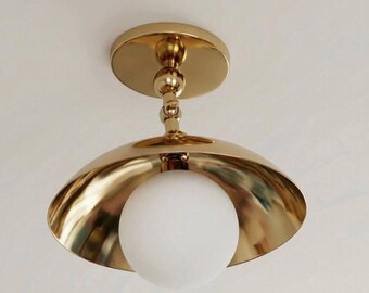 May include: A gold-colored ceiling light fixture with a spherical white glass shade. The fixture has a rounded, bowl-shaped shade and a circular base. The light is mounted on a white ceiling.