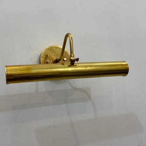 Könnte beinhalten: Eine Messing-Bildleuchte mit runder Wandhalterung, gebogenem Arm und zylindrischem Leuchtengehäuse. Die Leuchte besteht aus glänzendem Messing und ist zur Beleuchtung von Kunstwerken oder anderen Wandobjekten konzipiert. Sie ist an einer weißen Wand montiert.