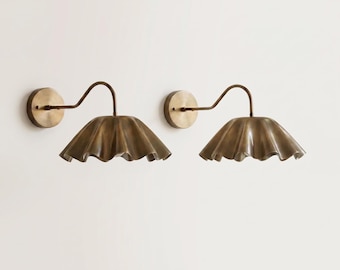 May include: Two brass wall sconces with a unique, ruffled shade design. Each sconce features a circular wall mount, a curved arm, and a shade with a wavy, textured edge. The sconces are mounted on a white wall, creating a minimalist aesthetic.