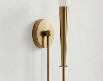 May include: A wall-mounted light fixture featuring a circular brass backplate and a slender, U-shaped arm that supports a vertical, tapered brass cone. A small, spherical white glass bulb sits at the top of the cone, completing the structure.
