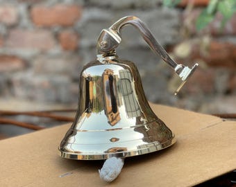 May include: A polished brass bell with a curved handle and clapper. The bell has a wide base and a smooth, reflective surface. The handle is attached to the top of the bell and curves upward. The bell is sitting on a cardboard surface.