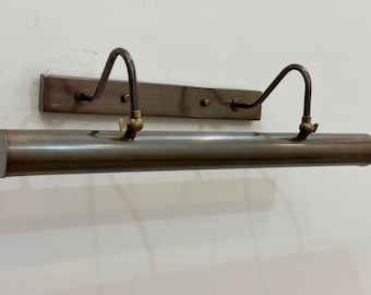 May include: A vintage-style picture light fixture with a brass finish. The fixture has a long, cylindrical light housing and two curved arms. It is mounted on a rectangular backplate with two small screws.
