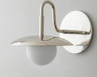May include: A modern wall sconce with a polished nickel finish. It features a circular backplate, a curved arm, and a rounded shade with a white globe light. The design is minimalist and elegant, suitable for various interior styles.