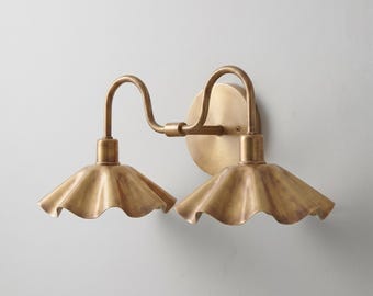 May include: A double wall sconce light fixture with a brushed brass finish. It features a circular wall mounting plate, two curved arms, and two wide, scalloped, bell-shaped shades that flare outward at the bottom edges, creating a symmetrical design.