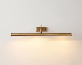 May include: A wall-mounted picture light with a long, cylindrical horizontal bar in a brushed brass finish. The fixture features a central rectangular mounting plate and a single vertical arm connecting the base to the light tube, emitting a warm glow.