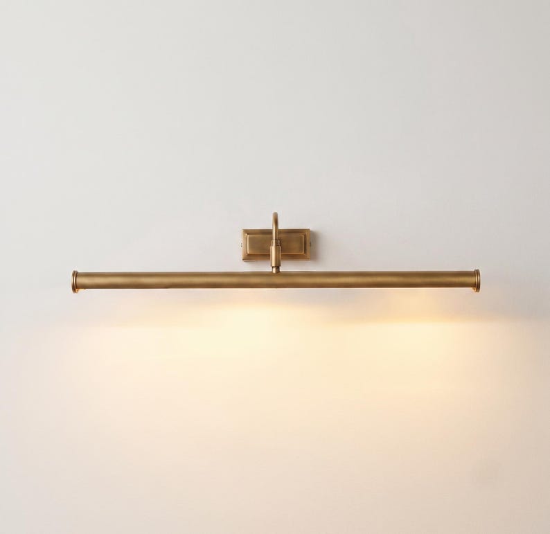 May include: A wall-mounted picture light with a long, cylindrical horizontal bar in a brushed brass finish. The fixture features a central rectangular mounting plate and a single vertical arm connecting the base to the light tube, emitting a warm glow.