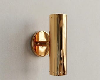 May include: A gold-colored wall sconce with a cylindrical shade and a circular base. The sconce has a polished finish and small perforations near the bottom of the shade. The fixture is mounted on a white wall.