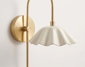 May include: A wall-mounted lamp featuring a brushed gold metal arm with a circular base and a downward-curving neck. The fixture includes a white, scalloped, bell-shaped shade that flares outward at the bottom, attached to the gold metal frame.