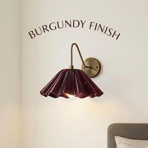 May include: A wall-mounted lamp featuring a burgundy finish, a pleated bell-shaped shade, and a curved brass-toned arm. The text BURGUNDY FINISH is printed above the fixture, which is attached to a circular wall plate with a single light bulb.