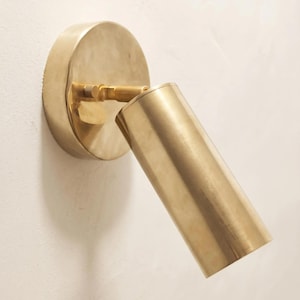 May include: A wall-mounted spotlight fixture crafted from brushed brass. The design features a circular base plate attached to a cylindrical light head, connected by a small adjustable joint with a textured knob for positioning the light angle.
