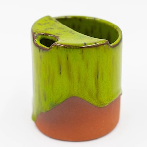 Könnte beinhalten: Eine Keramikvase mit einzigartigem Design. Die obere Hälfte ist leuchtend grün mit einem Tropfglasureffekt, während der Boden warmbraun ist. Die Vase hat eine gebogene Öffnung und ein kleines Loch in der Nähe der Oberseite.
