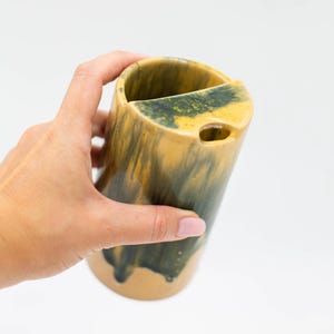 Könnte beinhalten: Eine zylindrische Keramikvase mit einem einzigartigen Design. Die Vase hat einen beigefarbenen Sockel mit einer dunkelgrünen und gelben Glasur, die an den Seiten herunterläuft. Sie hat einen Ausschnitt oben und ein kleines Loch an der Seite. Die Vase wird von einer Hand gehalten.
