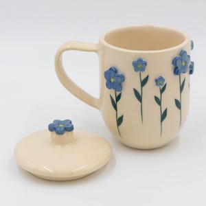 Könnte beinhalten: Cremefarbene Keramiktasse mit passendem Deckel. Die Tasse ist mit blauen Blumen und grünen Blättern verziert. Der Deckel hat einen blauen Blumenknauf. Die Tasse hat einen gebogenen Henkel.