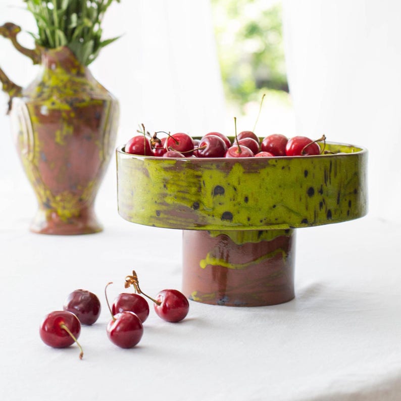 Handmade ceramic pedestal bowl from Mavroni Studio placed on a white linen tablecloth with soft natural light coming through sheer curtains. The bowl is empty, showcasing its glossy green glaze and red clay base. A ceramic vase with flowers is blurred in the background, and the Mavroni Studio logo is subtly displayed in the corner