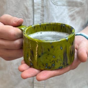 Può includere: Una tazza in ceramica fatta a mano con una vivace glassa verde e un design maculato. La tazza ha una forma cilindrica con un piccolo manico ed è riempita con una bevanda schiumosa. La tazza è tenuta tra le mani di qualcuno.