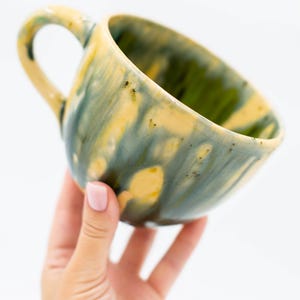 May include: A ceramic mug with a unique glaze. The mug features a handle and a rounded shape. The exterior is painted with a blend of yellow, green, and blue hues. The interior is green. The mug is held by a hand against a white background.