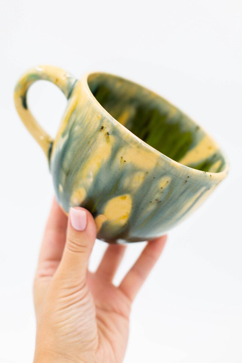 May include: A ceramic mug with a unique glaze. The mug features a handle and a rounded shape. The exterior is painted with a blend of yellow, green, and blue hues. The interior is green. The mug is held by a hand against a white background.