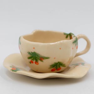 May include: A cream-colored ceramic teacup and saucer set. The cup has a unique, wavy rim and a small handle. Both pieces are decorated with green sprigs, red berries, and small orange dots. The saucer has a matching wavy edge.