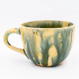 May include: A handmade ceramic mug with a rounded shape and a handle. The mug features a blend of colors, including yellow, green, and blue, with a dripped glaze effect. The interior is a light yellow color.