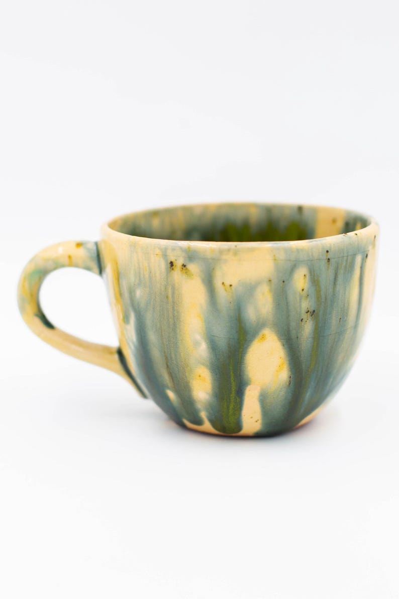 May include: A handmade ceramic mug with a rounded shape and a handle. The mug features a blend of colors, including yellow, green, and blue, with a dripped glaze effect. The interior is a light yellow color.