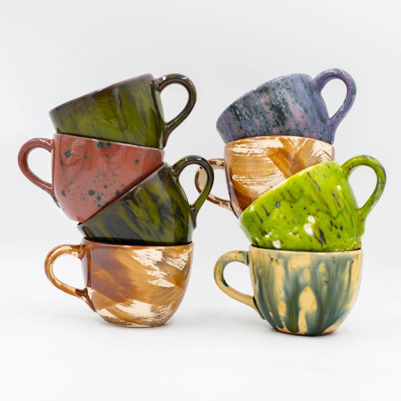 May include: A stack of handmade ceramic mugs in various colors, including green, brown, and purple. Each mug has a unique glaze and a curved handle. The mugs are stacked on top of each other, showcasing their different designs.
