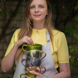 May include: A stack of four handmade ceramic mugs in shades of green, gray, and brown. The mugs have unique, textured finishes and curved handles. The person in the image is wearing a yellow top and a gray apron.