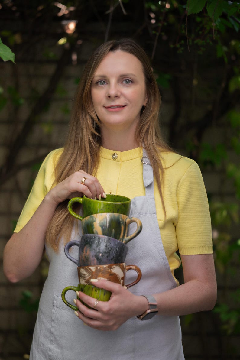 May include: A stack of four handmade ceramic mugs in shades of green, gray, and brown. The mugs have unique, textured finishes and curved handles. The person in the image is wearing a yellow top and a gray apron.