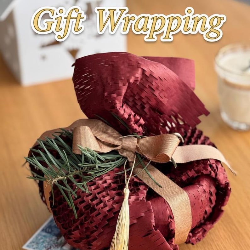May include: A wrapped gift with burgundy paper and a tan ribbon. The gift is decorated with a sprig of green pine needles and a gold tassel. The words "Gift Wrapping" are at the top of the image.