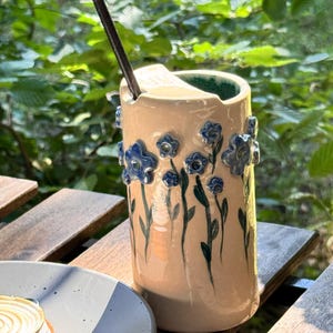 Puede incluir: Un vaso de cerámica beige con detalles florales azules y una pajita oscura. El vaso presenta flores azules pintadas a mano y tallos verdes. El borde interior es verde. El vaso está sobre una mesa de madera.