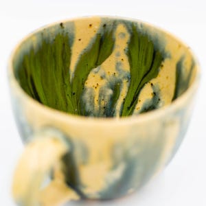 May include: A handmade ceramic mug with a unique design. The interior features a vibrant green and black swirl pattern, contrasting with the exterior's muted yellow and blue tones. The mug has a rounded shape with a comfortable handle, perfect for enjoying your favorite beverage.