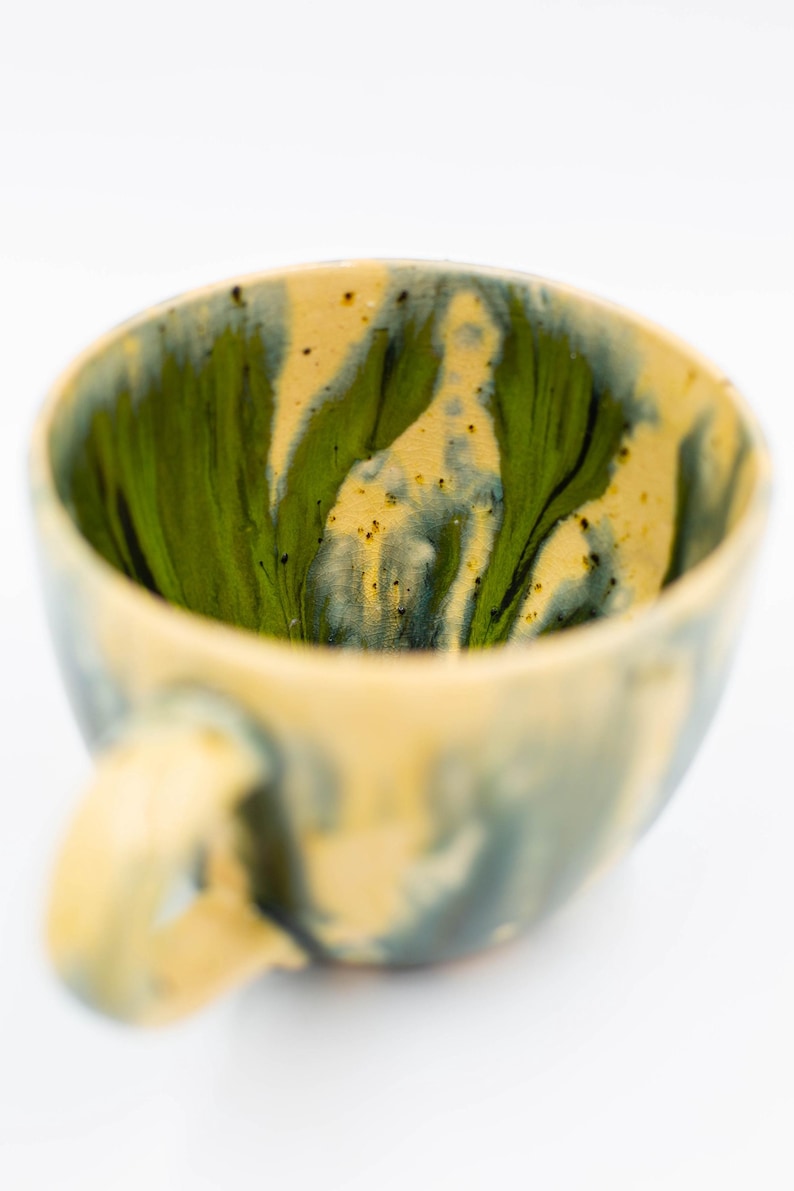May include: A handmade ceramic mug with a unique design. The interior features a vibrant green and black swirl pattern, contrasting with the exterior's muted yellow and blue tones. The mug has a rounded shape with a comfortable handle, perfect for enjoying your favorite beverage.