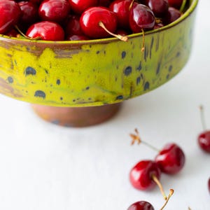 May include: A green ceramic bowl filled with ripe, red cherries. The bowl has a speckled design and a brown base. Several cherries are scattered on the white surface, showcasing their stems.