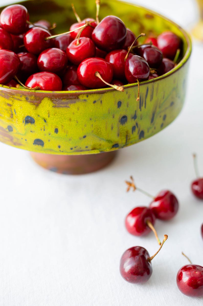 May include: A green ceramic bowl filled with ripe, red cherries. The bowl has a speckled design and a brown base. Several cherries are scattered on the white surface, showcasing their stems.