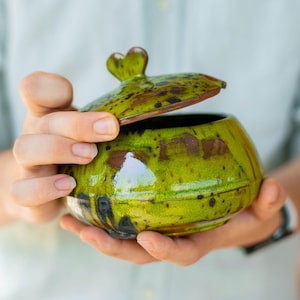 Puede incluir: Un azucarero de cerámica hecho a mano con tapa. El cuenco es de un verde vibrante con motas marrones y negras, y la tapa presenta un asa en forma de corazón. El cuenco se sostiene con ambas manos, mostrando su tamaño y diseño.