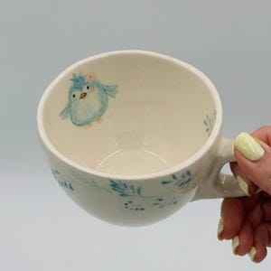 May include: Cream-colored ceramic mug with a whimsical blue bird illustration. The bird has a flower on its head. The mug features a handle and is decorated with teal floral accents around the rim. The mug is held by a person.