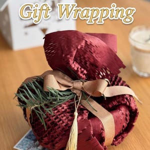 May include: A wrapped gift in burgundy paper, secured with a tan ribbon and bow. The gift is adorned with greenery and a gold tassel. The words "Gift Wrapping" are at the top of the image.