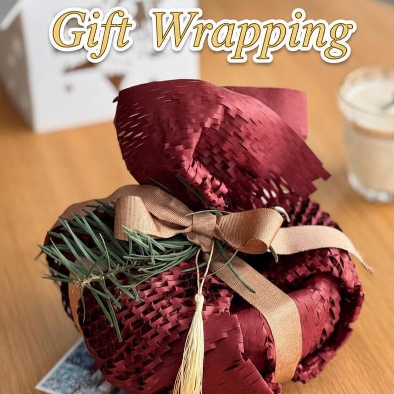 May include: A wrapped gift in burgundy paper, secured with a tan ribbon and bow. The gift is adorned with greenery and a gold tassel. The words "Gift Wrapping" are at the top of the image.
