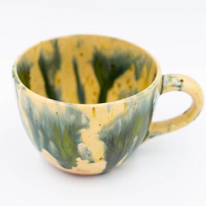 May include: A handmade ceramic mug with a unique glaze. The mug features a yellow base with green and blue flowing patterns. It has a rounded body and a curved handle, perfect for enjoying coffee or tea.