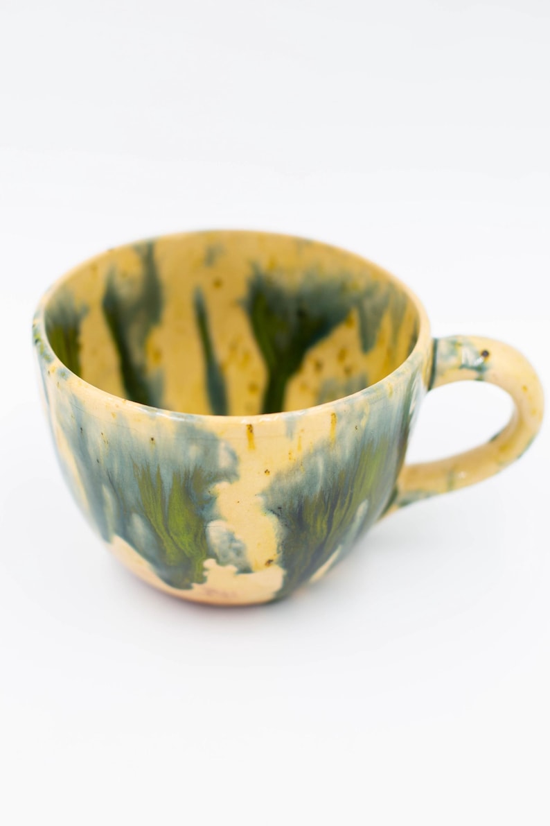 May include: A handmade ceramic mug with a unique glaze. The mug features a yellow base with green and blue flowing patterns. It has a rounded body and a curved handle, perfect for enjoying coffee or tea.