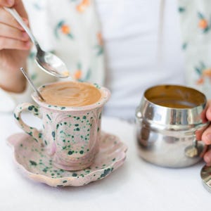 Puede incluir: Un juego de taza y platillo de cerámica rosa con motas verdes. La taza contiene café y una cucharita en su interior. Un azucarero plateado está junto a la taza. La persona lleva una camisa con estampado floral.