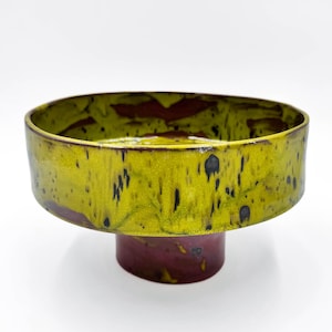May include: A ceramic pedestal bowl with a wide, flat rim. The rim is a vibrant yellow-green with black speckles, and the interior has a reddish-brown glaze. The pedestal base is a deep burgundy colour.