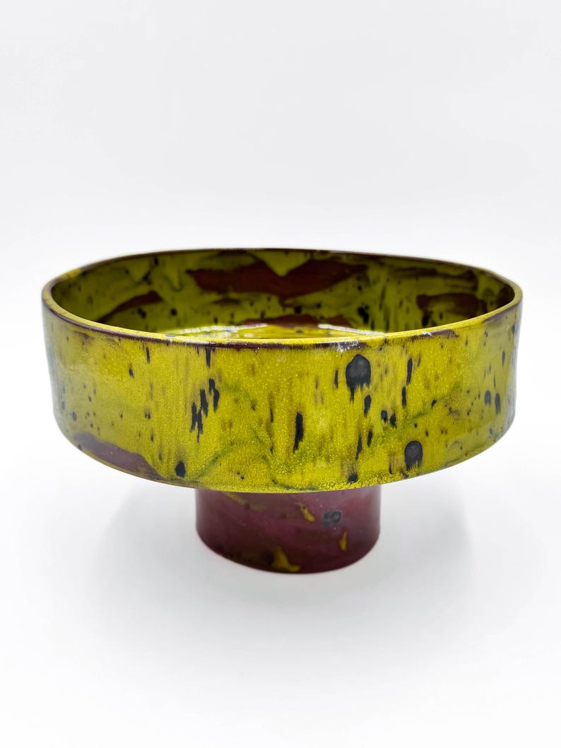 May include: A ceramic pedestal bowl with a wide, flat rim. The rim is a vibrant yellow-green with black speckles, and the interior has a reddish-brown glaze. The pedestal base is a deep burgundy colour.