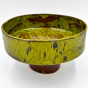 May include: A ceramic pedestal bowl with a wide, shallow design. The bowl has a vibrant yellow-green glaze with dark brown accents and speckles. The base is a dark brown colour, creating a striking contrast.