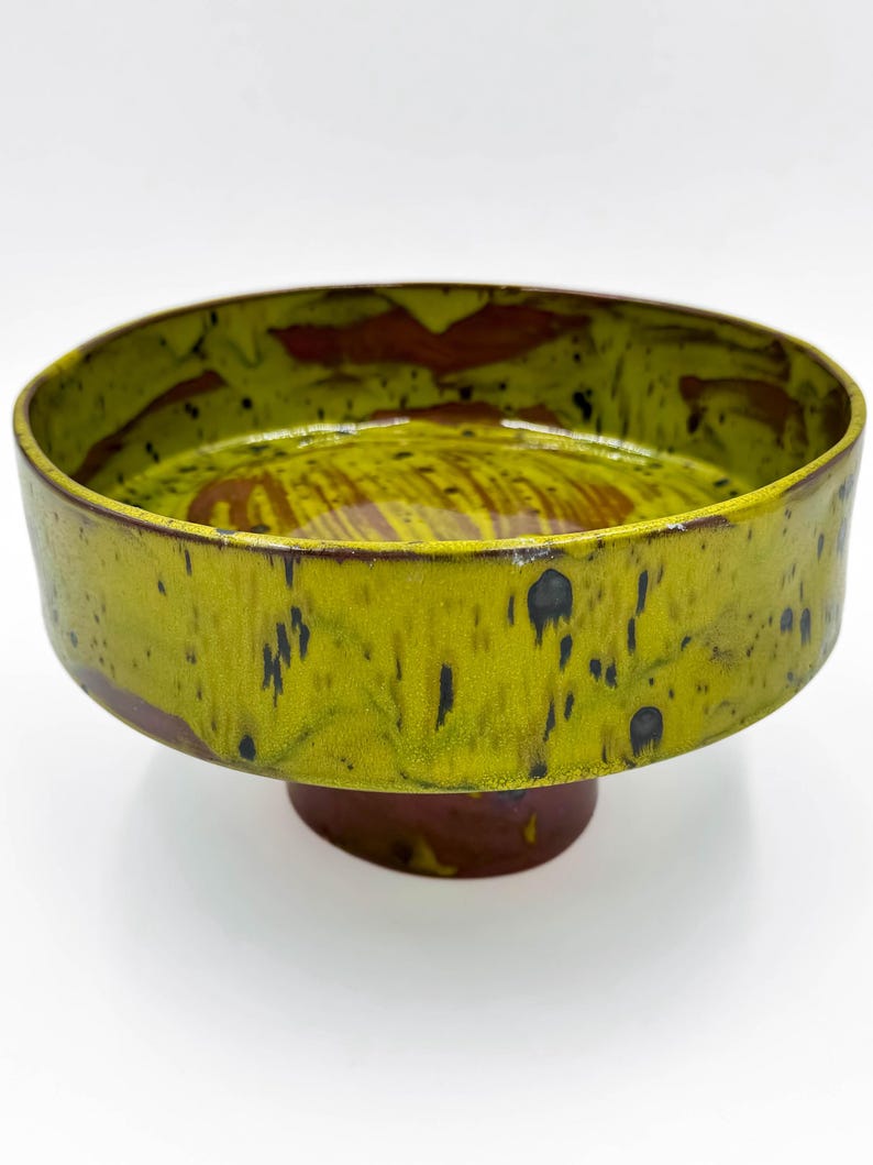 May include: A ceramic pedestal bowl with a wide, shallow design. The bowl has a vibrant yellow-green glaze with dark brown accents and speckles. The base is a dark brown colour, creating a striking contrast.