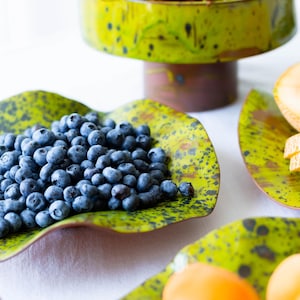 May include: A still life arrangement featuring fresh fruit on decorative serving dishes. A tiered pedestal bowl holds cherries, while blueberries and other fruits are displayed on green, speckled plates. The dishes have a unique, organic shape.
