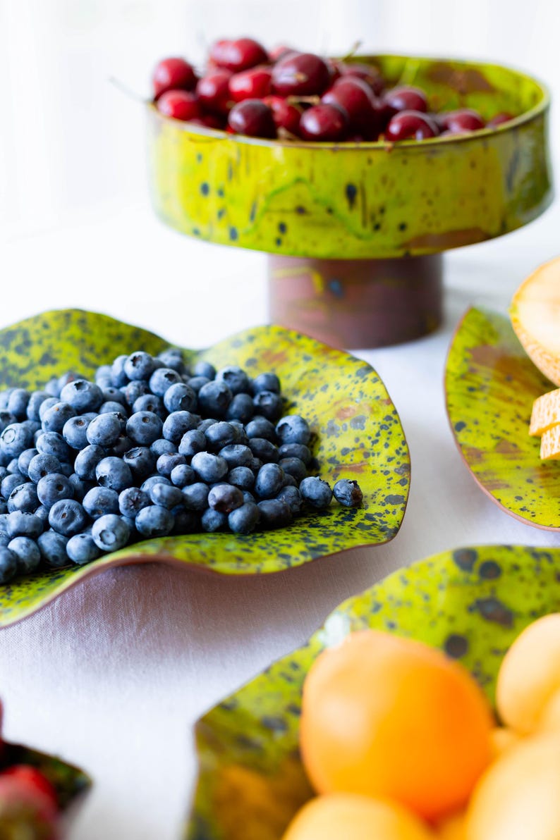 May include: A still life arrangement featuring fresh fruit on decorative serving dishes. A tiered pedestal bowl holds cherries, while blueberries and other fruits are displayed on green, speckled plates. The dishes have a unique, organic shape.