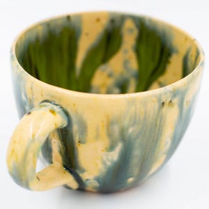 May include: Handmade ceramic mug with a unique glaze. The interior is a vibrant green, while the exterior features a blend of yellow, blue, and green hues. The mug has a rounded shape with a sturdy handle, perfect for enjoying your favorite beverage.